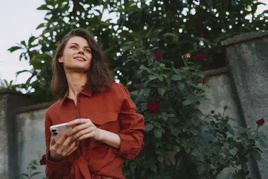 Улыбающаяся темноволосая женщина с телефоном на фоне розовых кустов