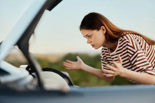 Раздражённая женщина в полосатой футболке жестикулирует у открытого капота машины