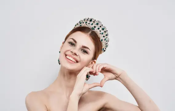 Улыбающаяся женщина в короне с изумрудами показывает сердечко руками