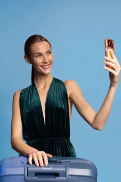 Молодая женщина в элегантном зеленом платье делает селфи на смартфон, держа в руке синий чемодан