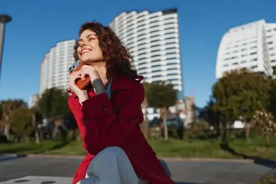 Улыбающаяся кудрявая девушка в красном пальто на фоне высоток