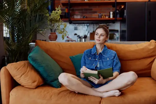 Молодая женщина в джинсовой куртке читает книгу на оранжевом диване