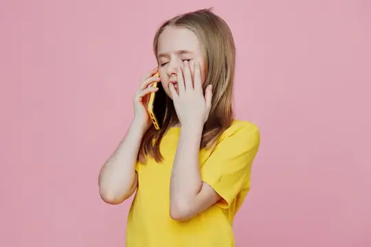 Юная девушка в жёлтой футболке, проявляющая эмоции во время разговора по телефону на фоне ярко-розового фона