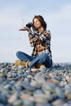 Молодая женщина с фотокамерой сидит на галечном пляже и подбрасывает камешки