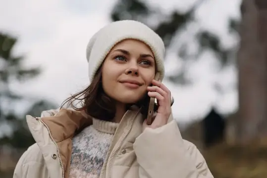Повседневная женская фигура в пуховике и белой шапке-бини разговаривает по мобильному телефону на улице, с туманным небом и естественным фоном, уличная сцена, передающая общение, моду и повседневную…