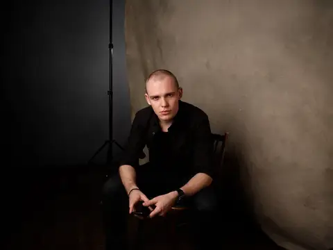 Молодой бритоголовый мужчина в чёрной рубашке сидит на стуле в фотозоне