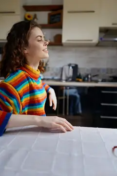 Молодая женщина в ярком свитере мечтательно улыбается на кухне