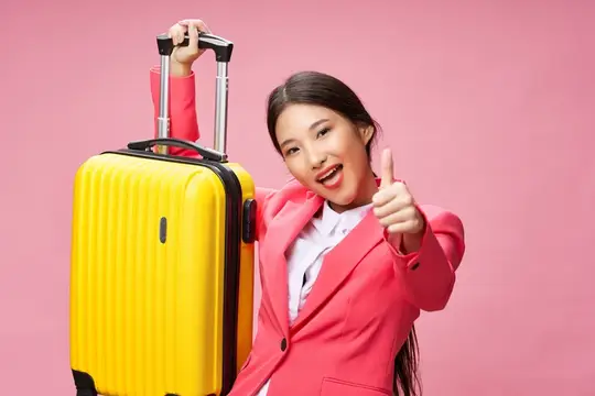 Улыбающаяся азиатская девушка с желтым чемоданом показывает большой палец вверх