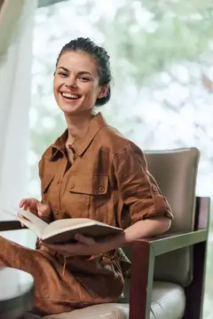 Улыбающаяся темноволосая женщина с книгой в кресле у окна