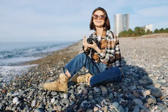 Портрет женщины-фотографа на открытом воздухе на каменистом берегу, держит камеру, одета в джинсы и фланель, наслаждается теплым солнечным светом и морским бризом, документируя сцены природы и…