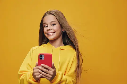 Улыбчивая девочка в жёлтом худи держит красный смартфон на жёлтом фоне
