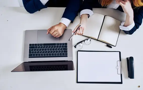 Двое коллег в деловых костюмах работают за столом с ноутбуком и блокнотом