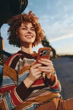 Улыбающаяся женщина с кудрявыми волосами держит смартфон на улице, в полосатом свитере, на обочине дороги на закате, повседневный момент, тёплый золотистый свет, повседневная вязаная мода, общение