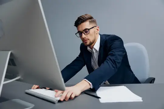Молодой мужчина в очках и деловом костюме работает за компьютером в офисе