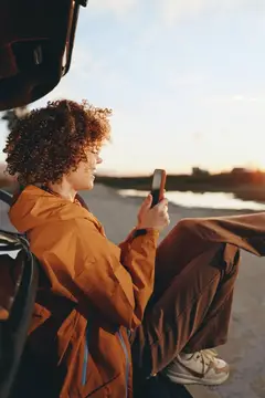 Кудрявый парень в оранжевой куртке смотрит в телефон на закате