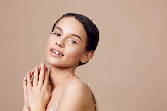 Молодая женщина с улыбкой касается лица руками на бежевом фоне