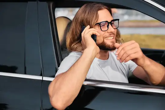Улыбающийся длинноволосый мужчина в очках говорит по телефону из окна автомобиля