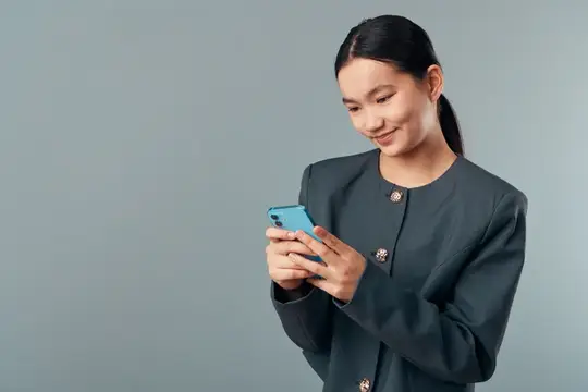 Улыбающаяся азиатская девушка в сером жакете смотрит в смартфон