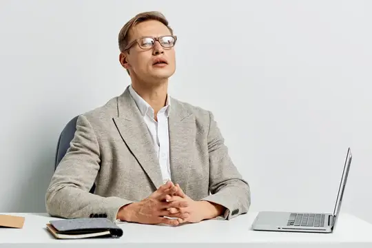Молодой мужчина в очках и сером пиджаке сидит за столом с ноутбуком