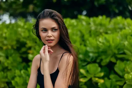 Молодая темноволосая женщина в чёрном топе на фоне зелёных кустов