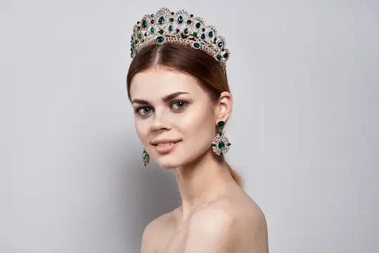 Молодая женщина в роскошной короне с изумрудами на сером фоне