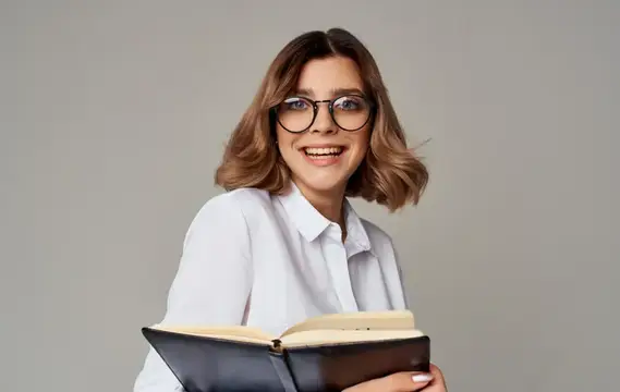 Улыбающаяся шатенка в очках с открытой книгой на сером фоне