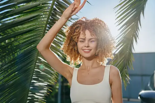 Улыбающаяся кудрявая девушка под пальмовыми листьями на солнце