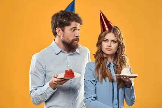 Мужчина и женщина в праздничных колпаках с грустными лицами держат маленькие торты