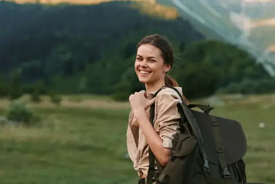 Улыбающаяся женщина с рюкзаком стоит на травянистом поле возле леса и гор, воплощая активный образ жизни и путешествия на свежем воздухе в естественной среде