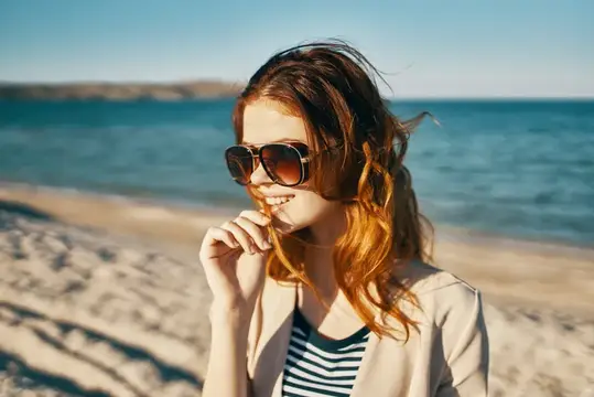 Счастливая рыжеволосая женщина в бежевой куртке и футболке стоит на песке у моря в горах