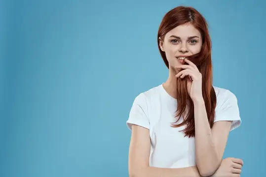 Рыжеволосая девушка в белой футболке игриво улыбается на голубом фоне