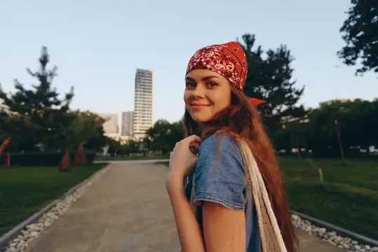 Женщина в стиле лайфстайл, одетая в современный бохо-вестерн деним стритстайл, уверенно улыбается на городской дорожке на фоне зелени