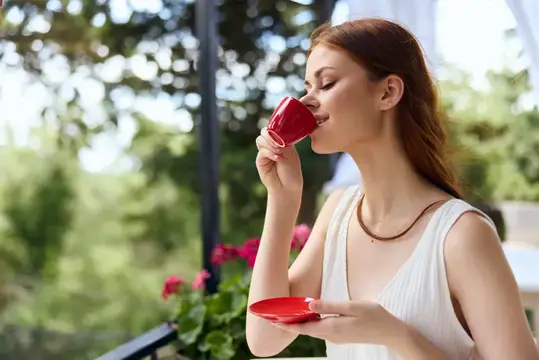 Рыжеволосая женщина с наслаждением пьёт из красной чашки на балконе