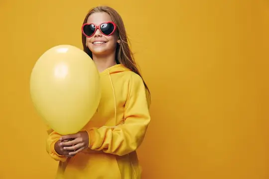 Улыбчивая девочка в красных очках держит жёлтый шарик на жёлтом фоне