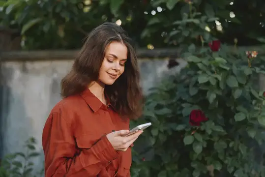 Молодая женщина с телефоном улыбается на фоне розовых кустов