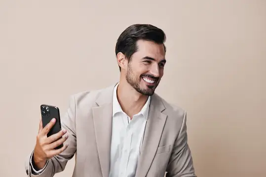 Улыбающийся бородатый мужчина в бежевом пиджаке смотрит на смартфон