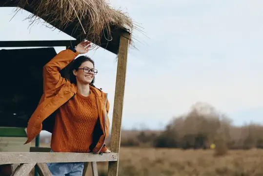 Улыбающаяся женщина в очках и оранжевом свитере стоит у деревянной постройки