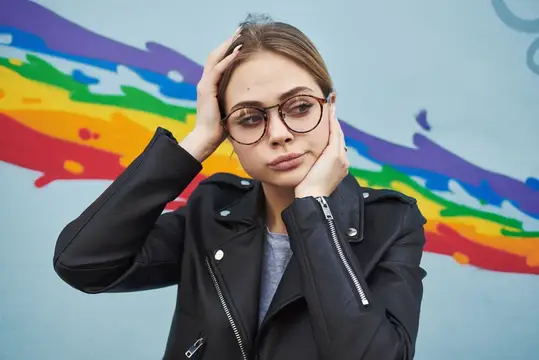 Молодая девушка в очках и черной косухе на фоне разноцветной стены