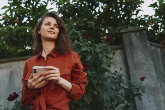 Повседневная уличная сцена: улыбающаяся женщина с телефоном в руке среди зелени, роз и солнечного света