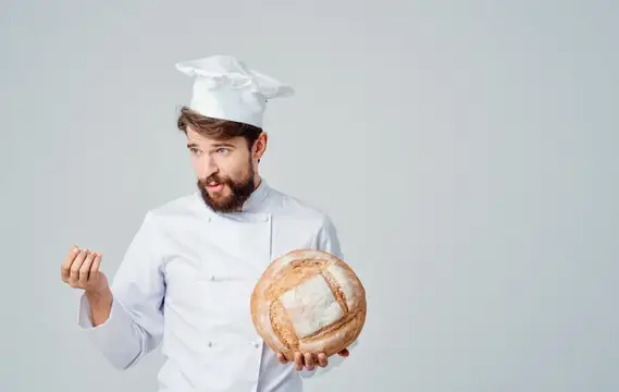 Шеф-повар в светлом костюме и головном уборе держит в руке круглый каравай хлеба