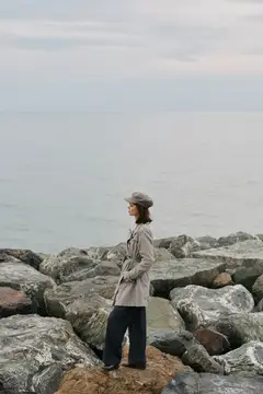 Девушка в плаще и шляпе стоит на камнях у моря в пасмурный день