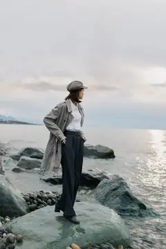 Женщина в плаще и кепке стоит на камнях у моря