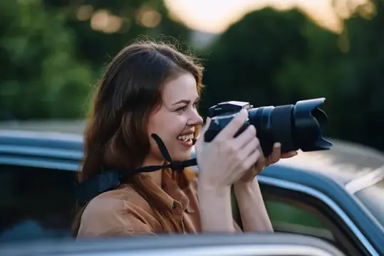 Улыбающаяся девушка с фотокамерой выглядывает из окна автомобиля