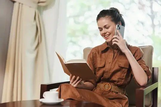 Улыбающаяся женщина с книгой говорит по телефону в кресле