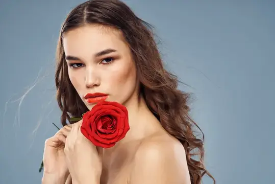 Портрет женщины на сером фоне: она держит красную розу у лица, у неё вьющиеся волосы и макияж