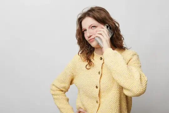 Кудрявая женщина в жёлтом вязаном свитере прижимает смартфон к уху с задумчивым видом