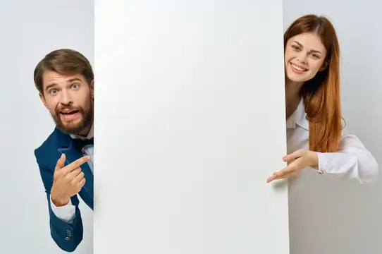 Удивлённый мужчина в костюме и улыбающаяся женщина выглядывают из-за белого баннера