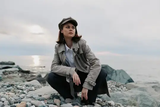Молодая женщина в кепке и плаще сидит на камнях у моря