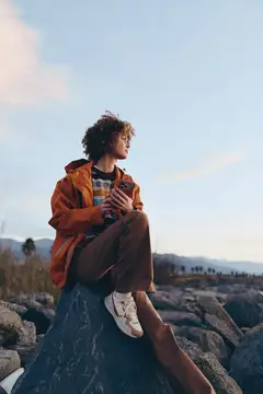 Кудрявый парень в оранжевой куртке сидит на камне в горах