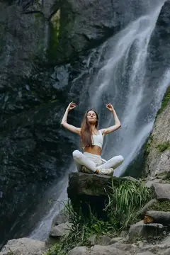 Молодая женщина медитирует в позе лотоса на камне у водопада